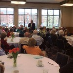 A man stands and gestures among tables of seated listeners
